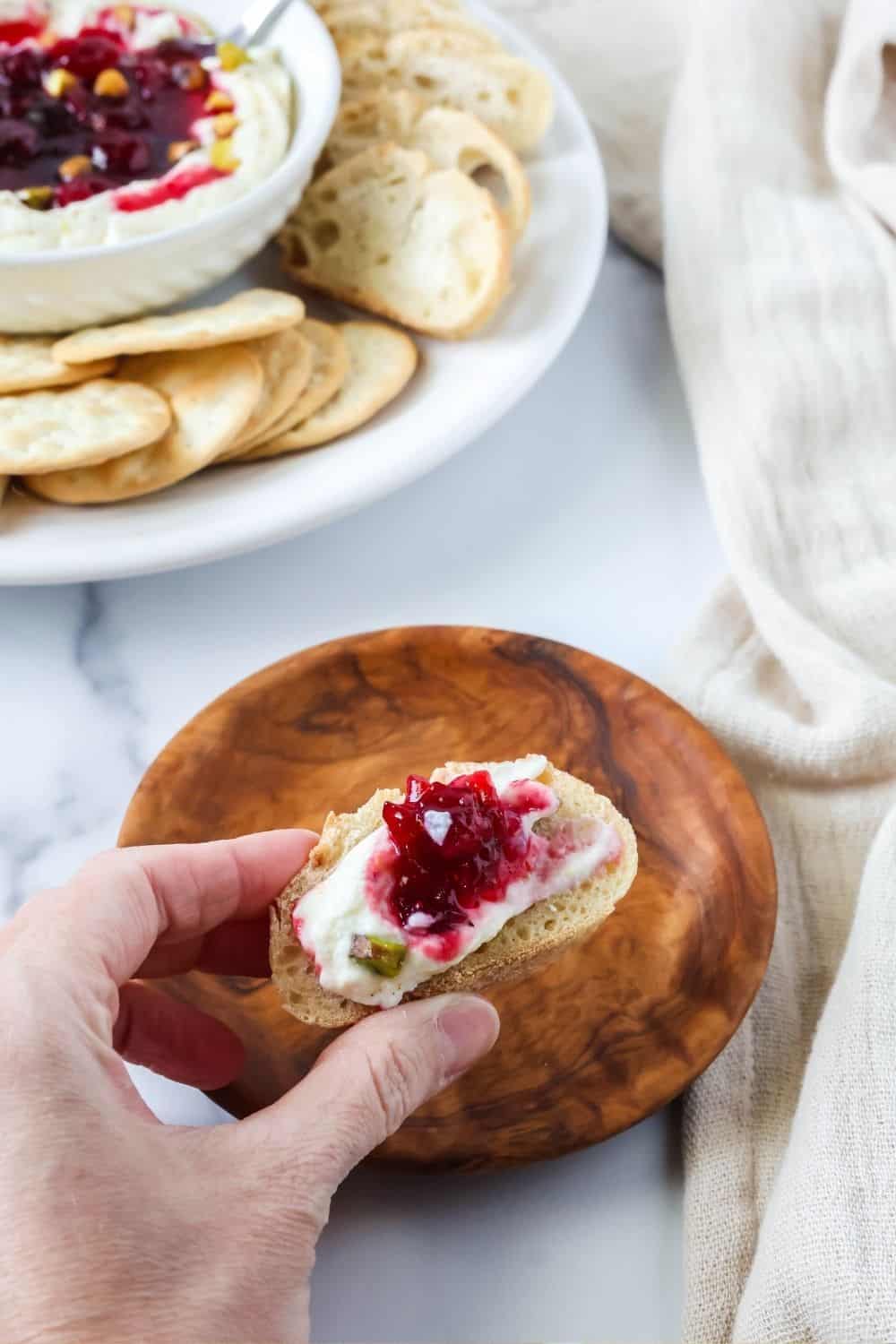 a hand holds a slice of baguette spread with whipped ricotta, cranberry sauce, and pistachios.