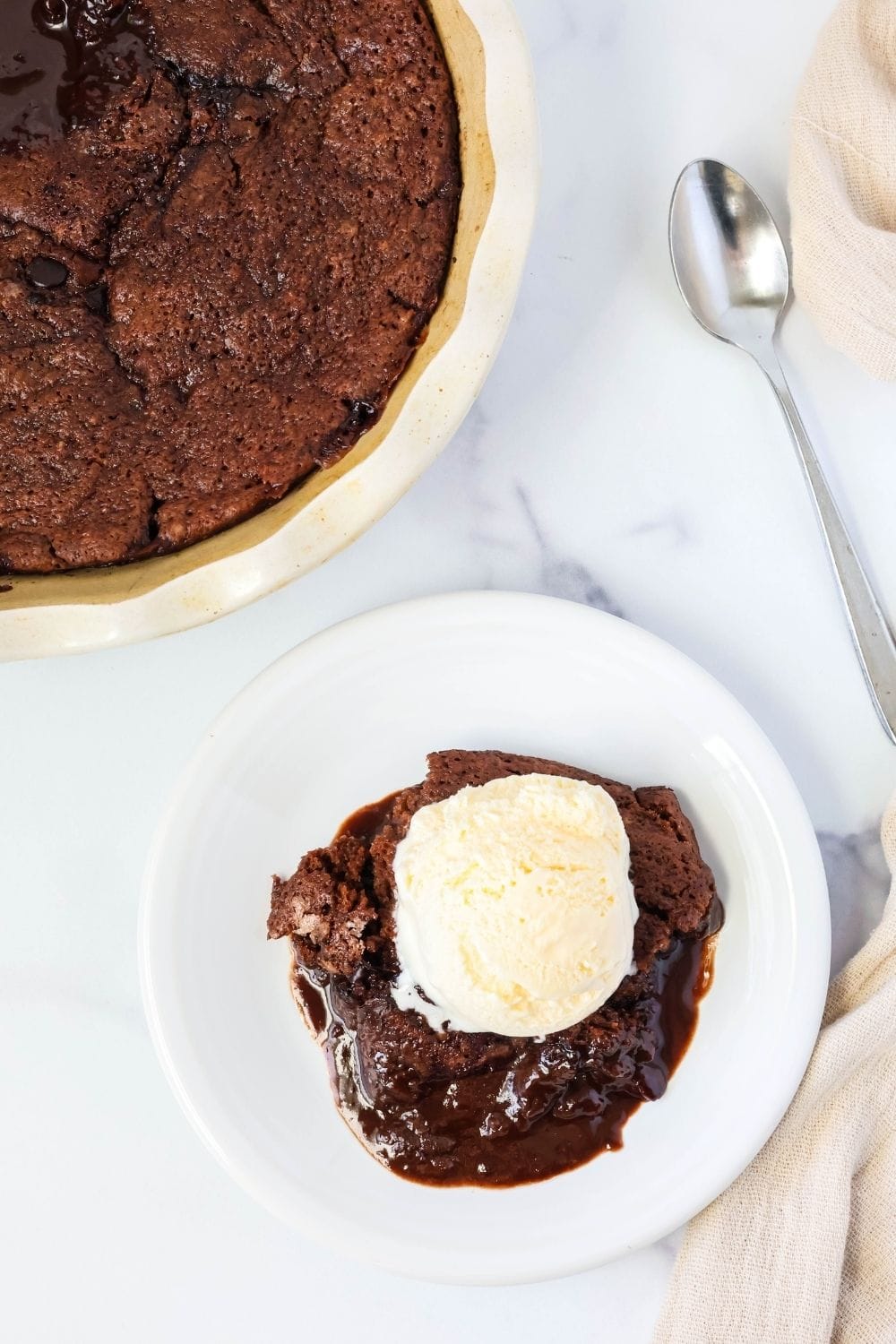 overhead view of a white plate with a serving of gooey chocolate cobbler topped with ice cream. The remaining dish of chocolate cobbler is in the corner, along with a spoon.