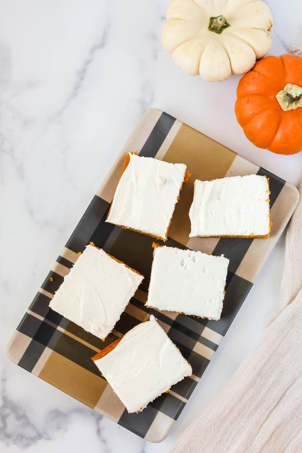 Overhead view of pumpkin cake squares topped with cream cheese frosting on a rectangular platter. Two mini pumpkins are next to the platter.