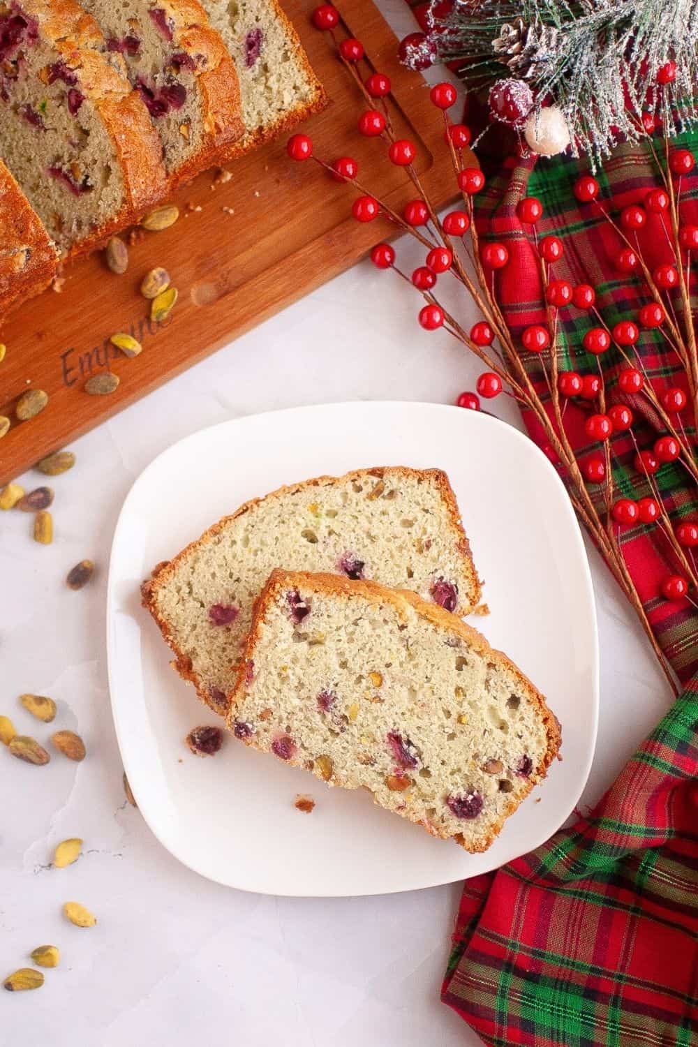 overhead view of two slices of pistachio bread with cranberries, served on a white plate. In the background is Christmas decor and a sliced loaf of cranberry pistachio bread on a cutting board.
