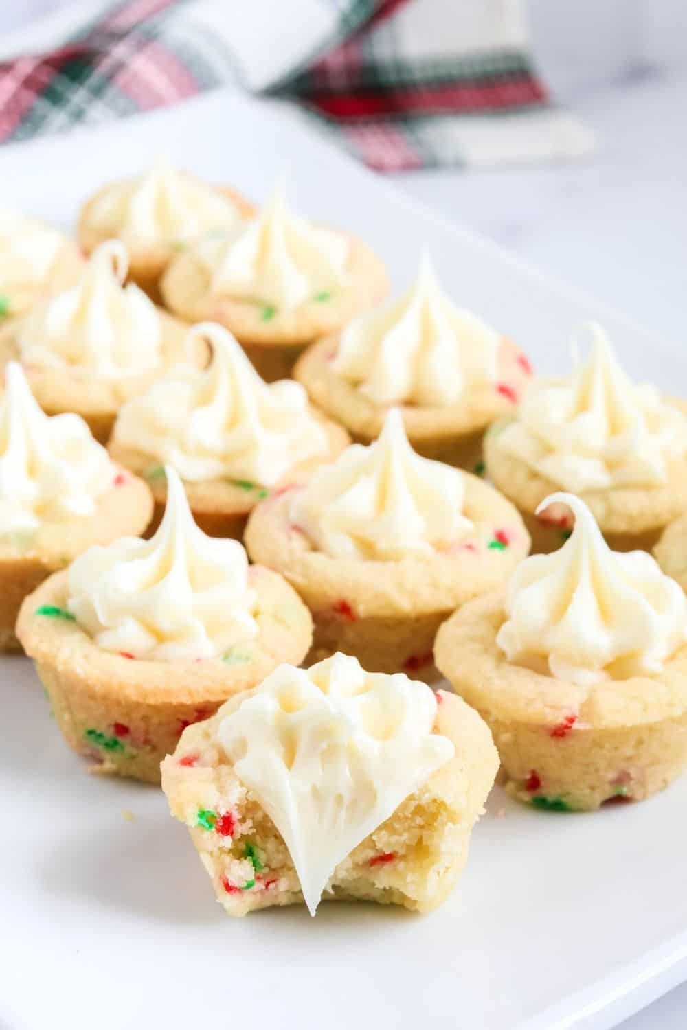 sugar cookie cups for Christmas displayed on a white platter; one of the cookie cups has a bite taken out of it, showcasing the soft sugar cookie exterior and the creamy vanilla frosting interior.