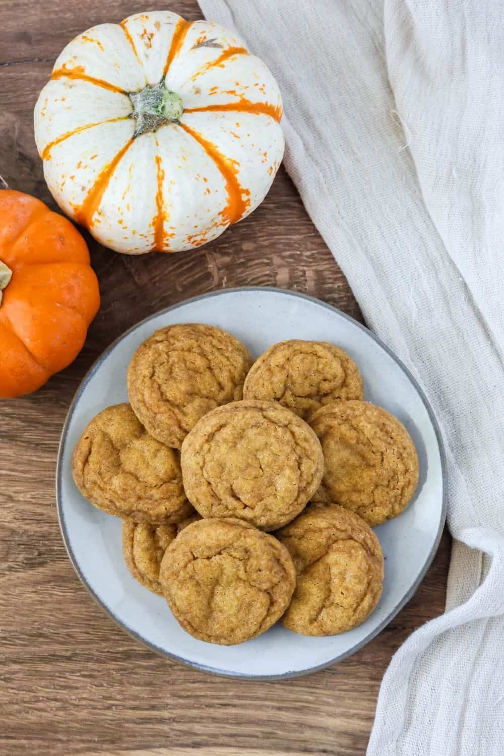 overhead view of chewy pumpkin snickerdoodle cookies on a cream-colored plate, with two miniature pumpkins next to it.