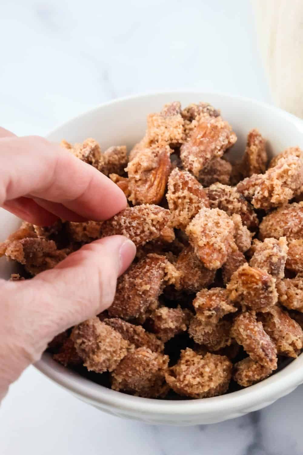 a woman's hand picks up a cinnamon sugar candied almond from a bowl of almonds.