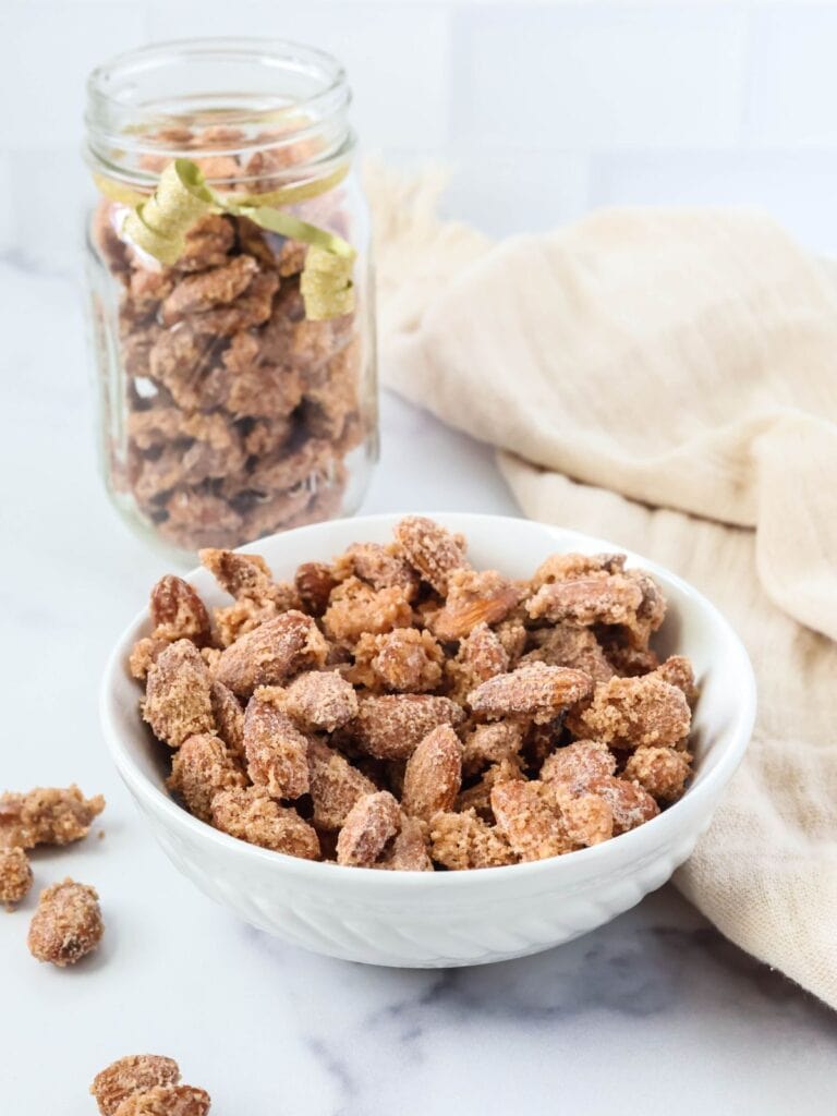 Stovetop cinnamon sugar candied almonds in a white bowl; with more candied almonds in a glass jar in the background.