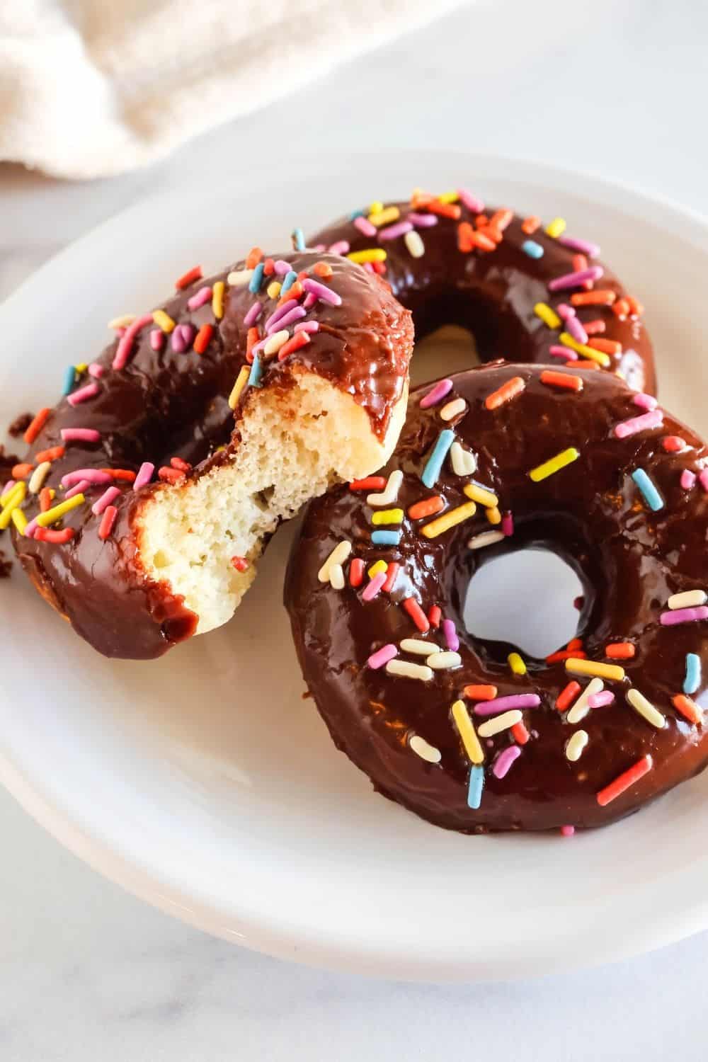 three baked donuts made with Bisquick, topped with chocolate glaze and sprinkles, served on a white plate. One of the donuts has a bite taken out of it, showing the soft and fulffy center of the donut.