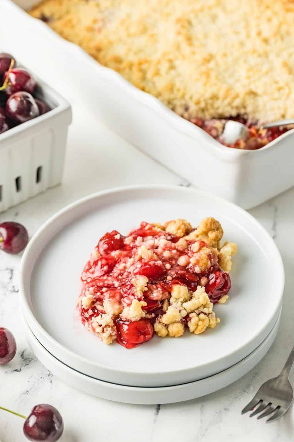 white plates with a serving of cherry cobbler made with pie filling and cake mix. In the background is the remaining baking dish of cobbler and a dish of cherries.