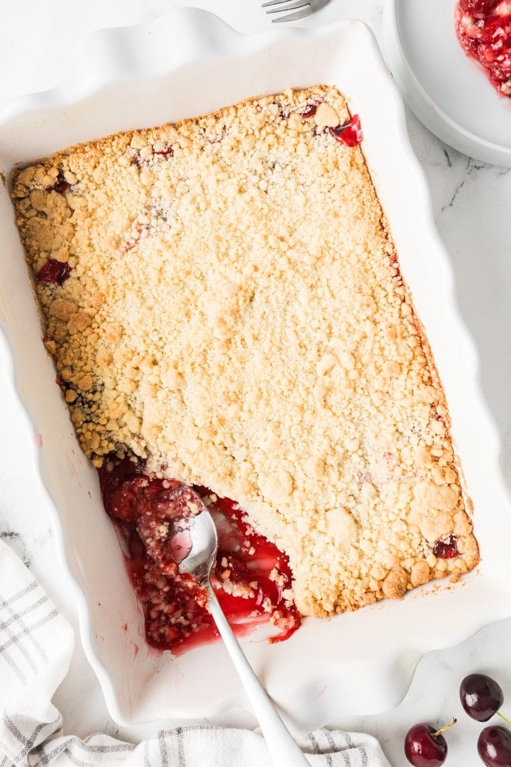 overhead angled view of a white baking dish with freshly baked cherry cobbler in it; a spoon is scooping out a serving of the cobbler.