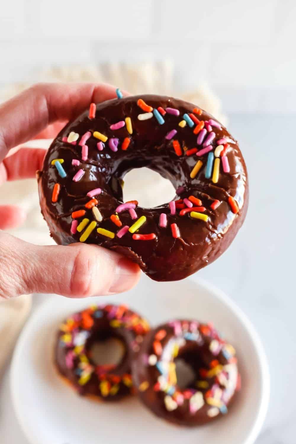 a hand holds a homemade baked donut topped with chocolate glaze and sprinkles.