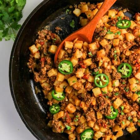 Mexican ground beef and potatoes, AKA picadillo con papas, in a cast iron skillet with a wooden spoon.
