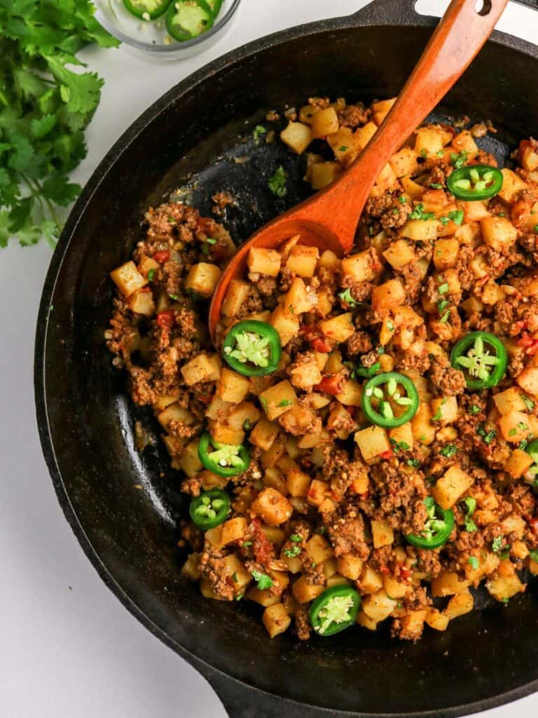 Mexican ground beef and potatoes, AKA picadillo con papas, in a cast iron skillet with a wooden spoon.