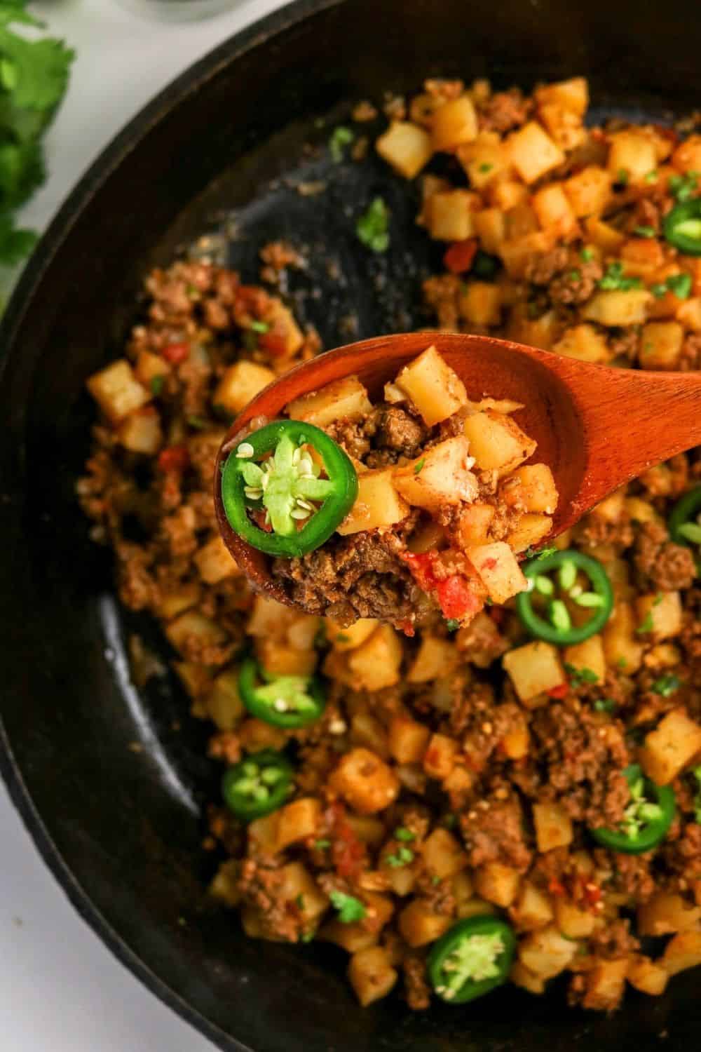 A wooden spoon scoops out Mexican ground beef and potatoes from a black skillet.