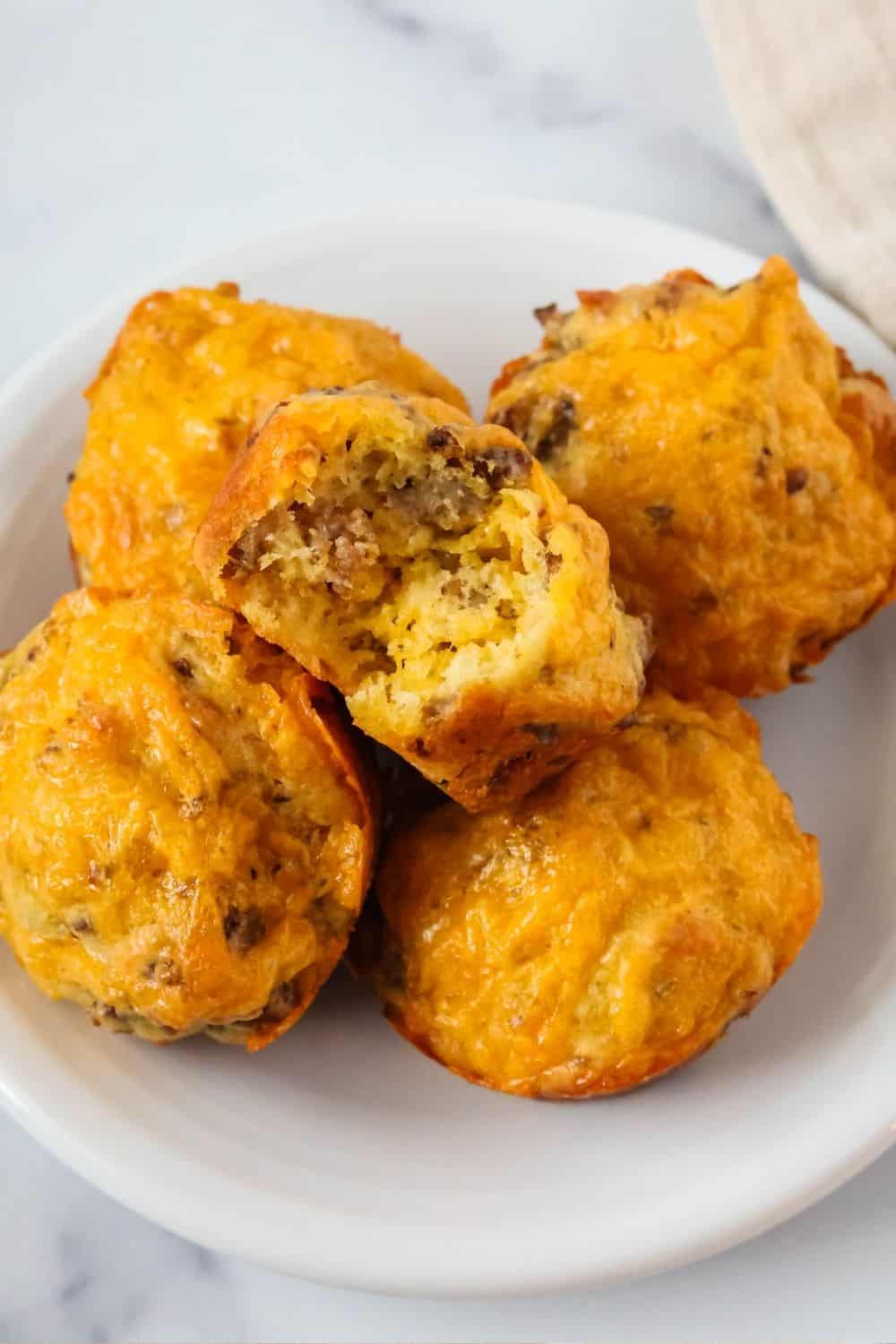 close-up view of several savory Bisquick muffins on a white plate. One muffin has a bite taken out of it, showing the cheese and sausage inside.