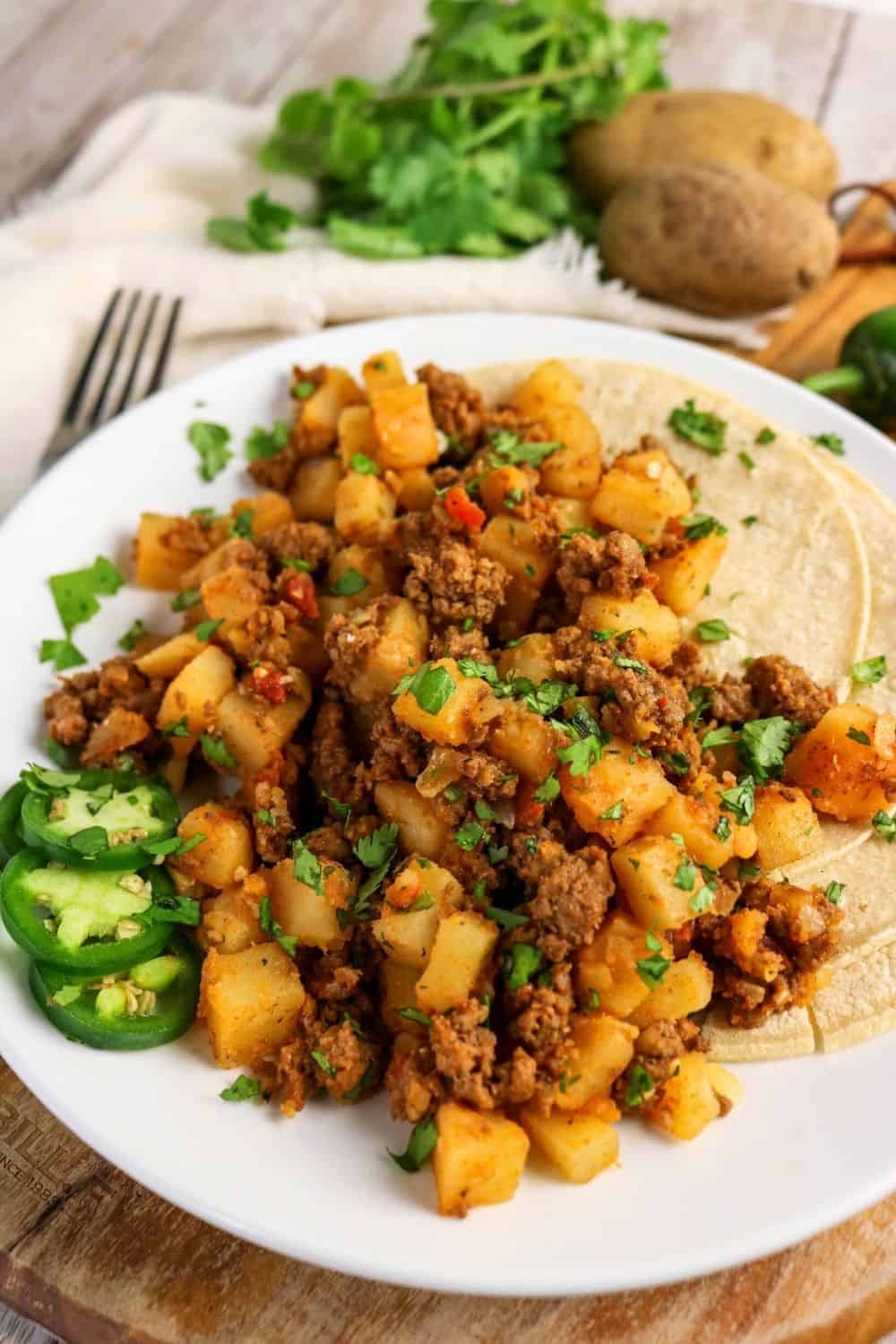 white plate with a serving of Mexican ground beef and potatoes alongside some corn tortillas, garnished with sliced jalapenos.