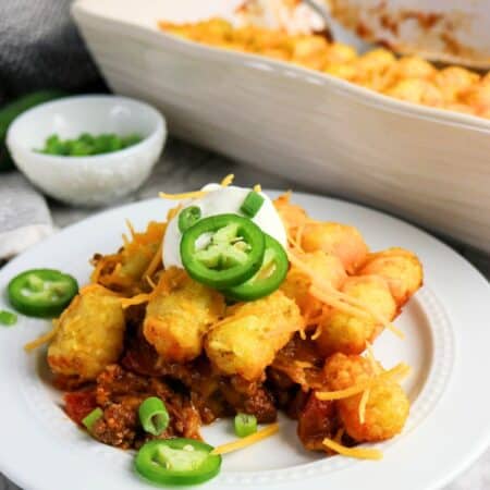 Chili tater tot casserole served on a white plate, with the casserole dish of remaining tater tot bake in the background.