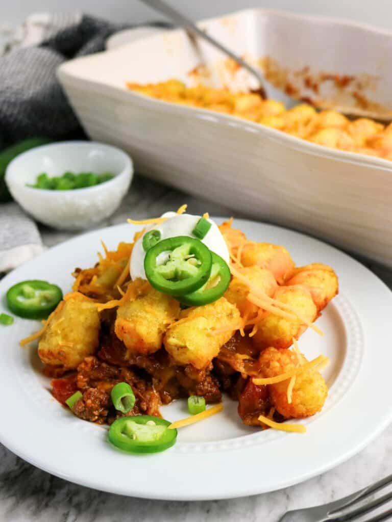 Chili tater tot casserole served on a white plate, with the casserole dish of remaining tater tot bake in the background.