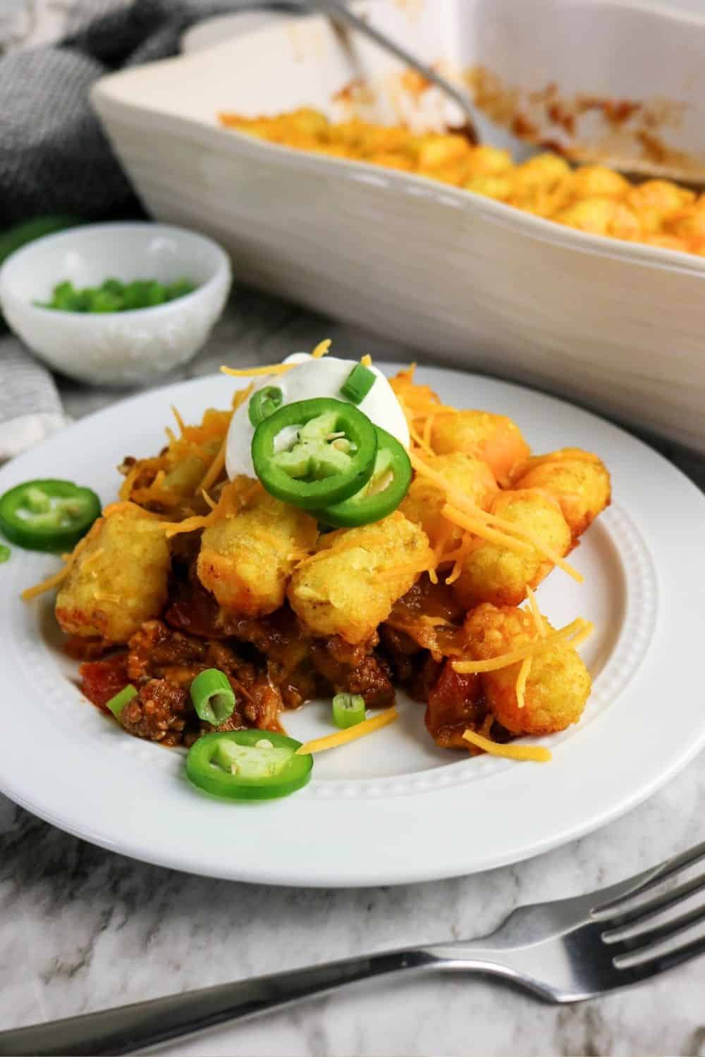 Mexican tater tot casserole on a white plate, with the baking dish of remaining casserole in the background.