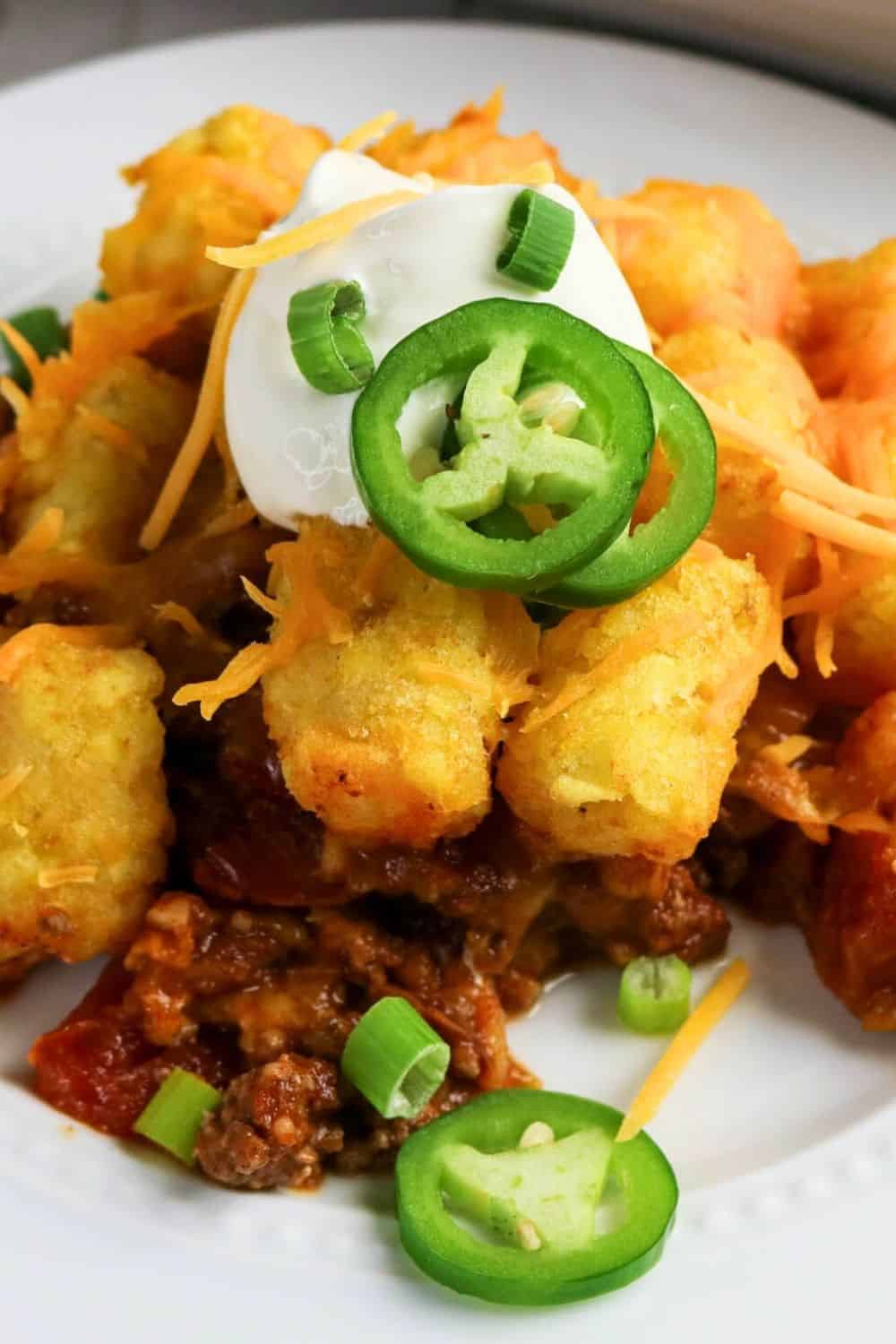close-up view of taco tater tot casserole on a white plate, garnished with green onions, jalapenos, and sour cream.