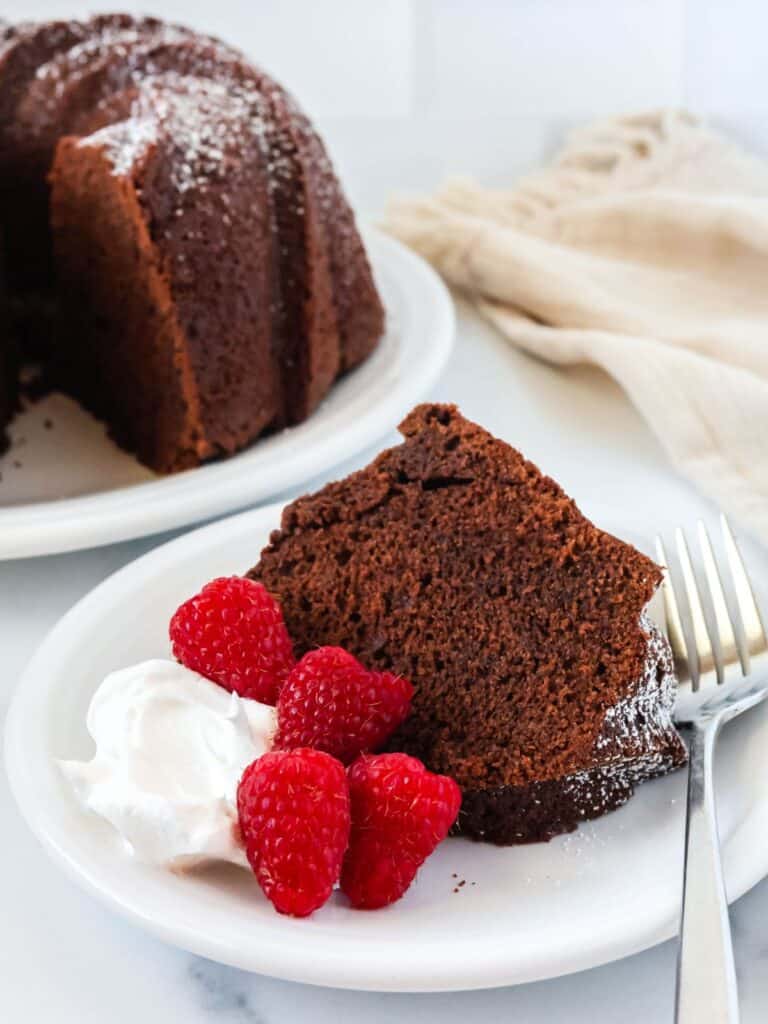 Slice of chocolate pound cake made from cake mix served on a white plate with fresh raspberries and whipped cream.