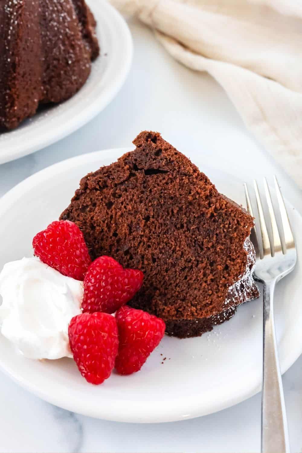 a slice of chocolate pound cake, raspberries, whipped cream, and a fork on a white plate.