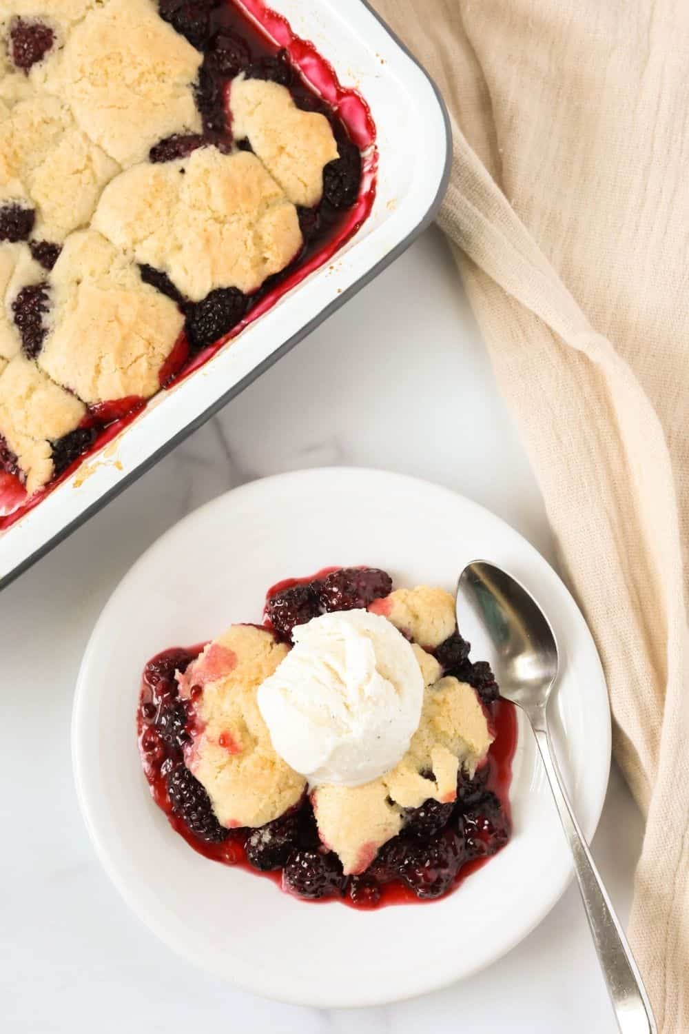 overhead view of a serving of blackberry cobbler with biscuit topping and a scoop of ice cream on a white dessert plate, next to the pan of freshly baked berry cobbler.