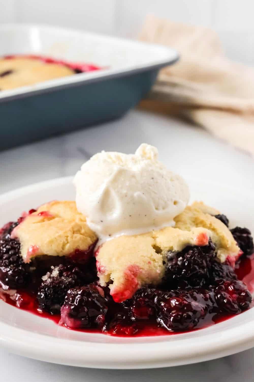 a plate of blackberry cobbler with Bisquick topping is in the foreground; the pan of cobbler is in the background.