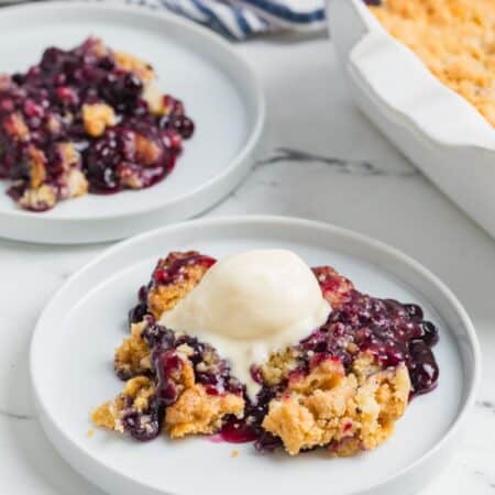 Blueberry cobbler dump cake served on white plates, with the baking dish of remaining cobbler in the background.