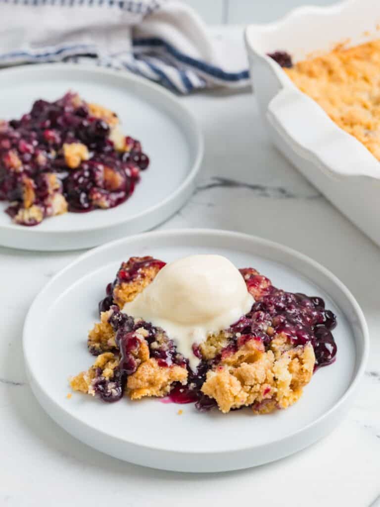 Blueberry cobbler dump cake served on white plates, with the baking dish of remaining cobbler in the background.