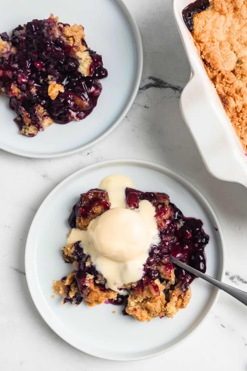 overhead view of two white plates with helpings of blueberry cobbler dump cake, one of which is topped with a scoop of vanilla ice cream.
