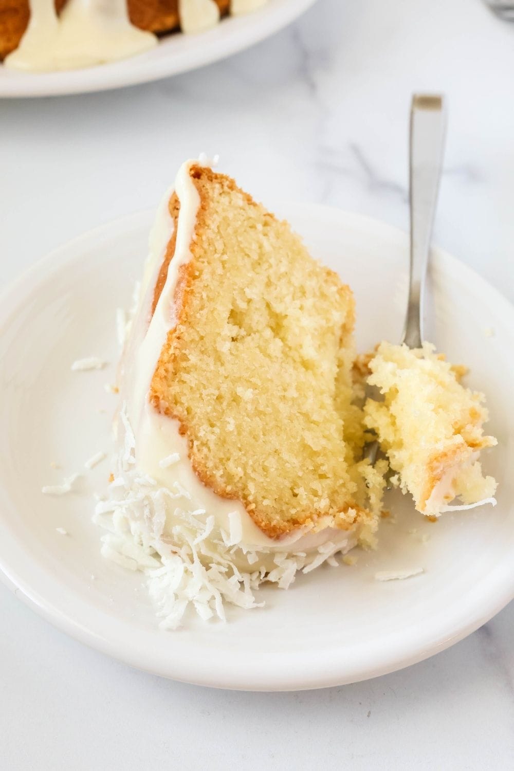 A slice of cake mix coconut pound cake topped with icing and shredded coconut, served on a white plate. A fork is cutting a bite out of the slice of cake.