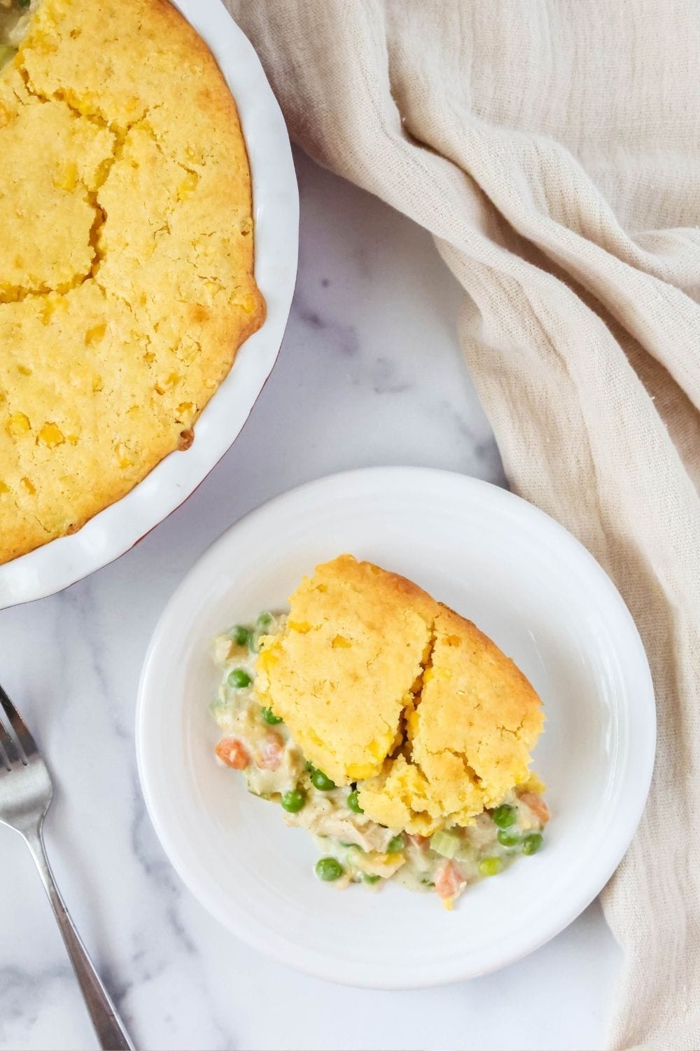overhead view of a small white plate with a helping of chicken pot pie cornbread casserole on it; the pie pan of remaining casserole is next to the plate.