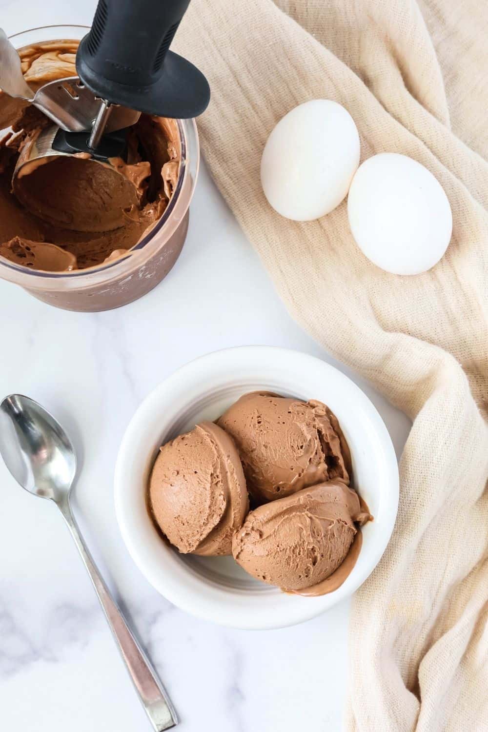 overhead view of a bowl of chocolate boiled egg ice cream scoops, with a pint container of remaining ice cream and two hard-boiled eggs in the background. A spoon is next to the bowl of ice cream.