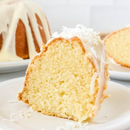 Slice of coconut pound cake served on a white plate. The remainder of the bundt cake and another slice of the cake are in the background.