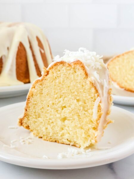 Slice of coconut pound cake served on a white plate. The remainder of the bundt cake and another slice of the cake are in the background.