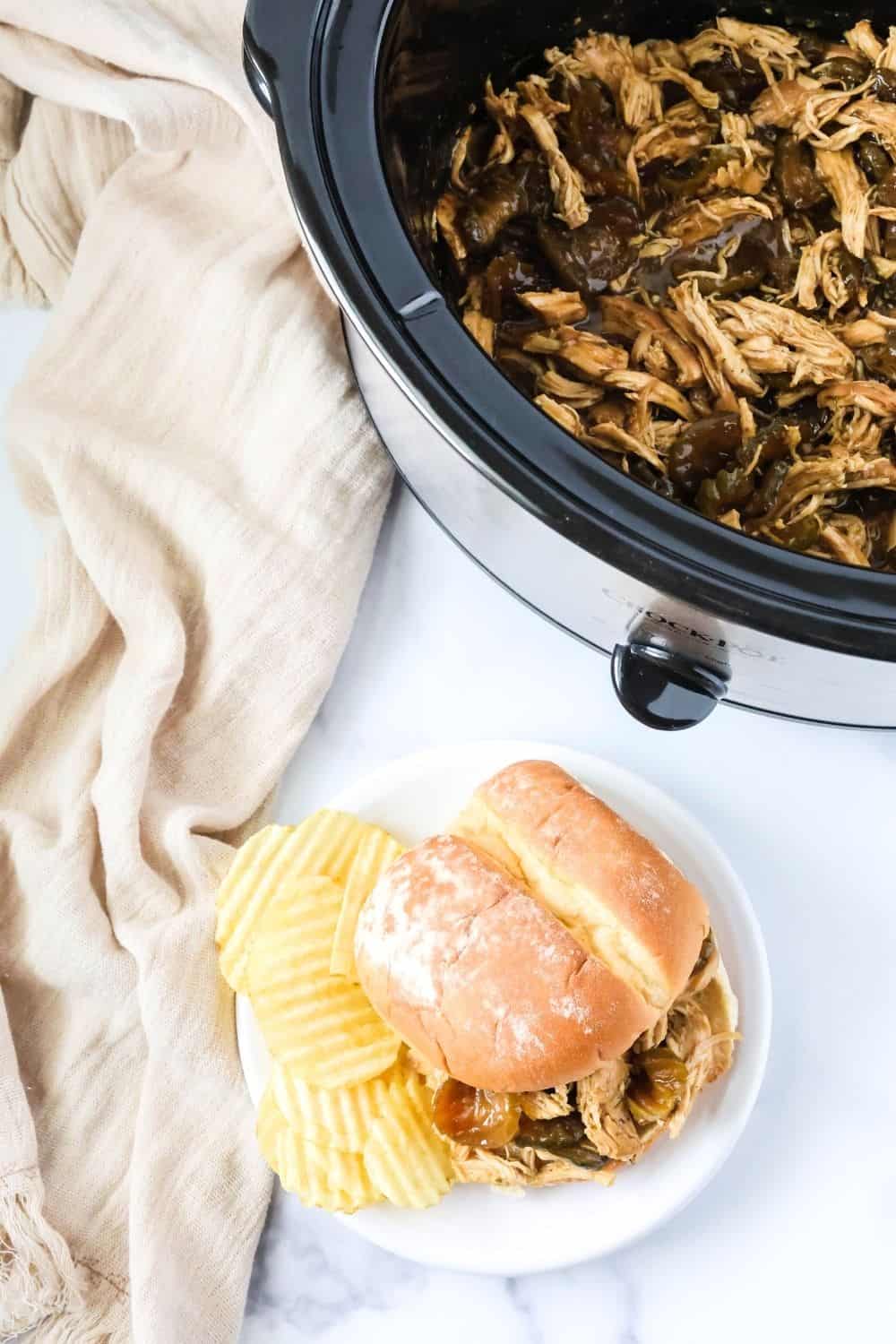 overhead view of a dill pickle chicken sandwich on a plate with potato chips, next to a Crock Pot filled with more of the shredded chicken and pickles.