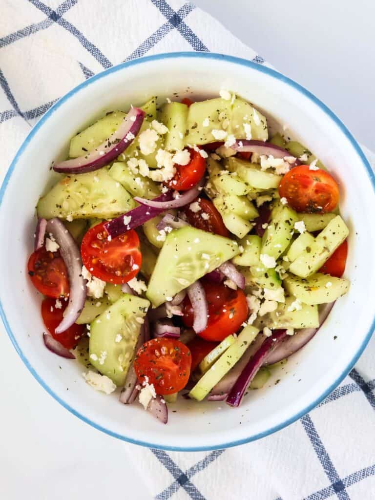 Cucumber salad with tomatoes, red onions, and feta in an herby vinaigrette, served in a white bowl with blue rim.