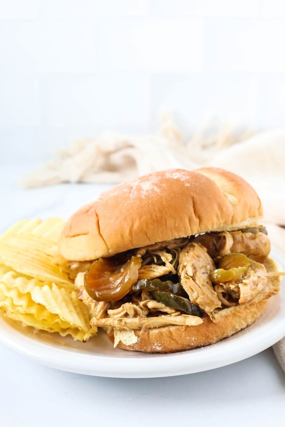 A bun filled with dill pickle chicken for sandwiches, served on a white plate with some potato chips.