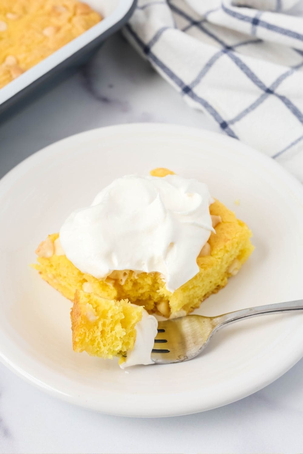 slice of lemon dump cake, topped with whipped cream, on a dessert plate. A fork has removed a bite of the cake.