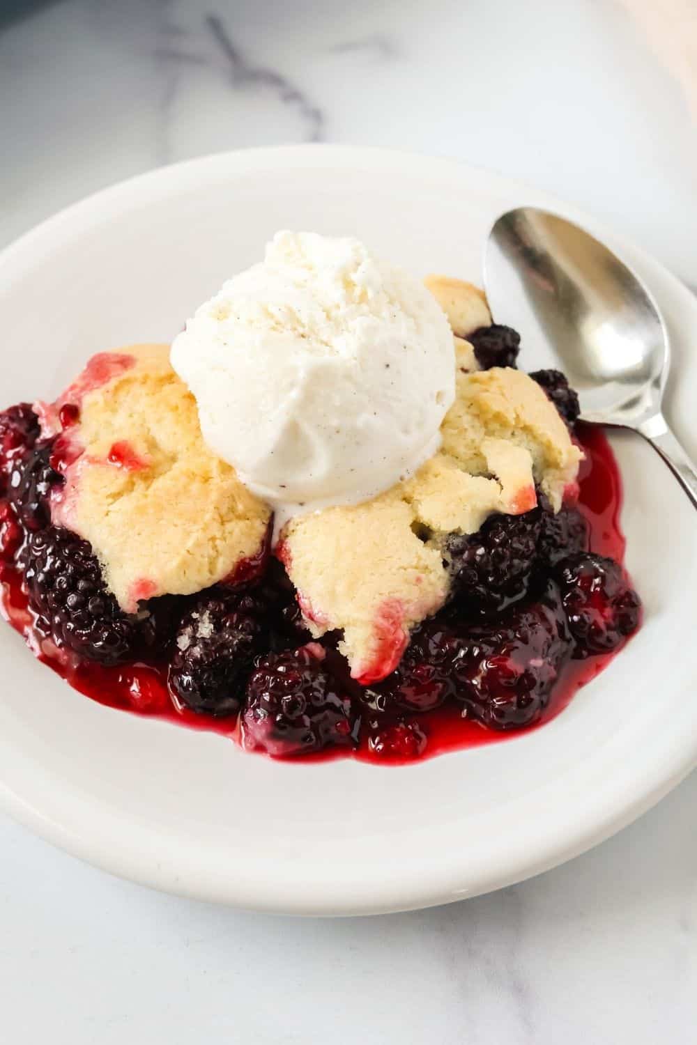 a serving of easy blackberry cobbler on a white plate, topped with vanilla ice cream. A spoon is on the plate.
