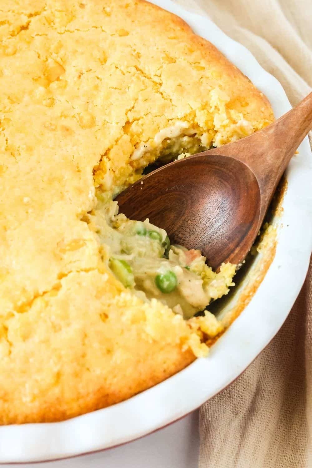 A wooden serving spoon scoops out a serving of homemade cornbread chicken pot pie casserole from a deep dish pie pan.