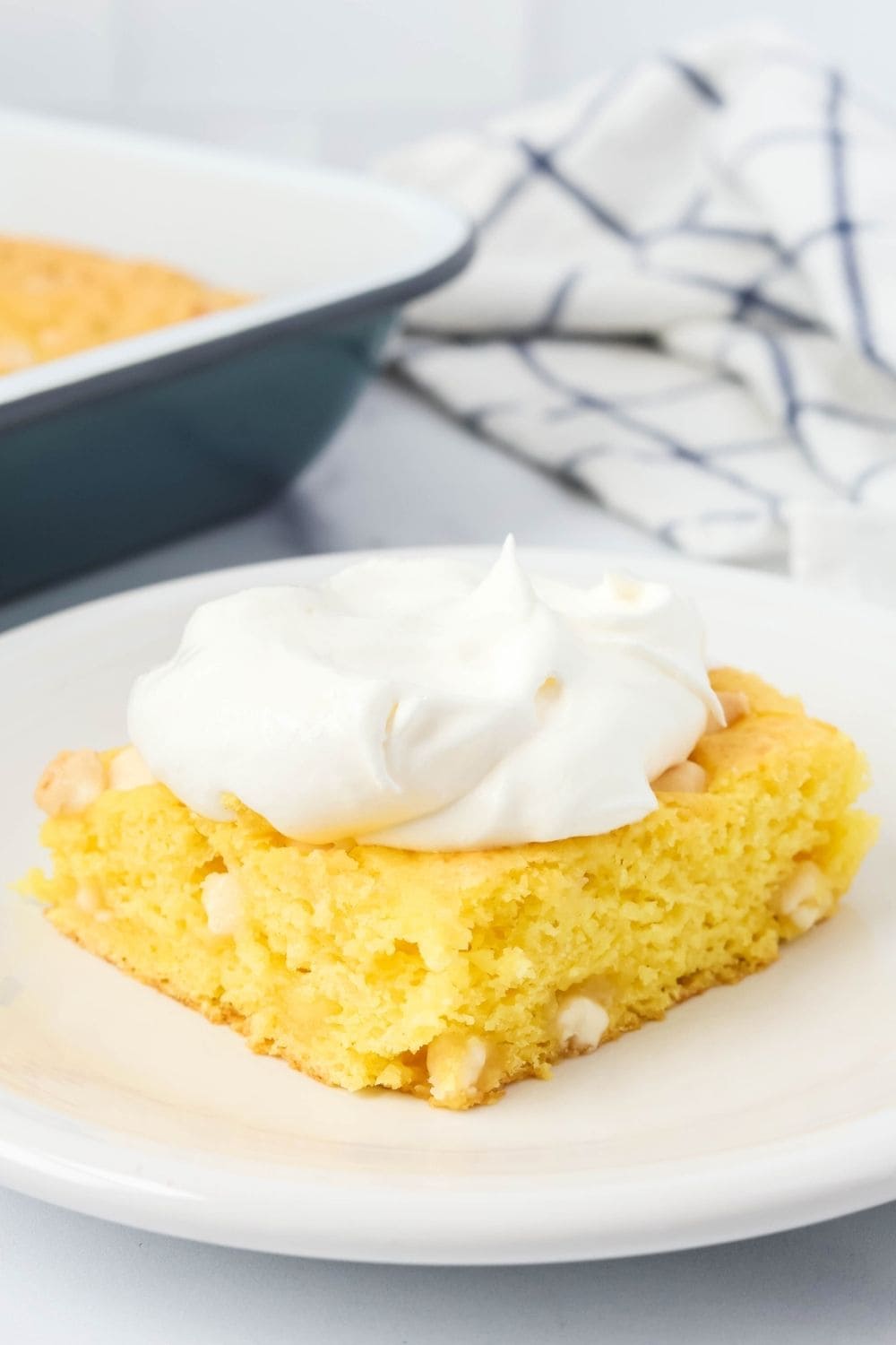 close-up view of lemon cake made without eggs or oil. The slice is topped with Cool Whip and served on a white plate. The cake pan is in the background.