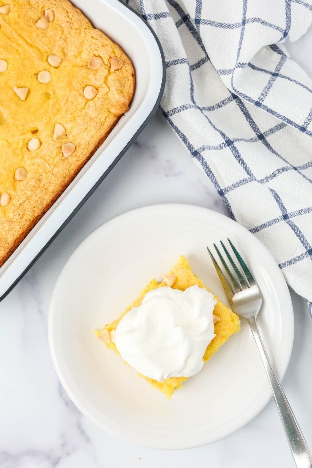 overhead view of a square of low fat lemon cake on a white plate, garnished with reduced-fat Cool Whip. A fork is on the plate, and the remaining pan of cake is next to it.