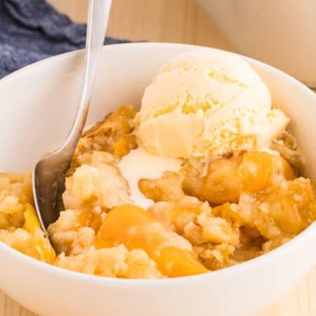 a serving of peach cobbler dump cake in a white bowl, topped with a scoop of vanilla ice cream. A spoon is in the bowl.