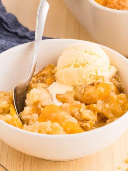 a serving of peach cobbler dump cake in a white bowl, topped with a scoop of vanilla ice cream. A spoon is in the bowl.