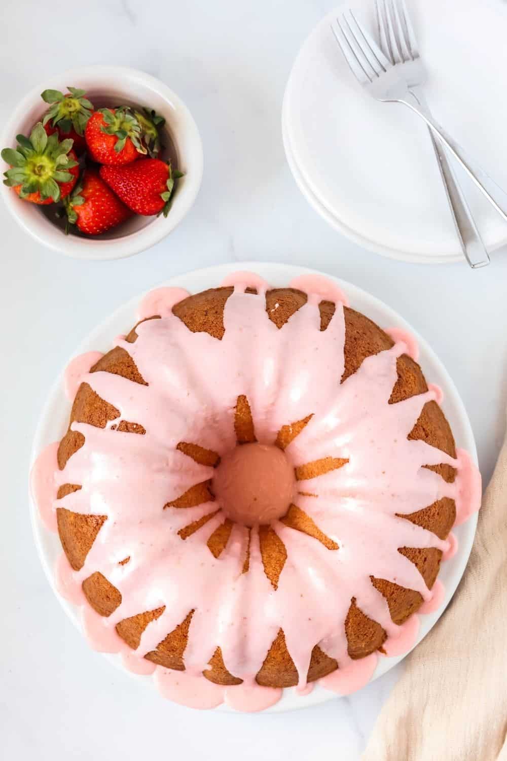 overhead view of a glazed strawberry cake mix bundt cake on a white plate, with fresh strawberries and small dessert plates in the background.