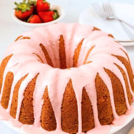 Strawberry pound cake made from a mix, topped with strawberry glaze, served on a white plate. A small bowl of fresh strawberries is in the background, along with dessert plates and forks.