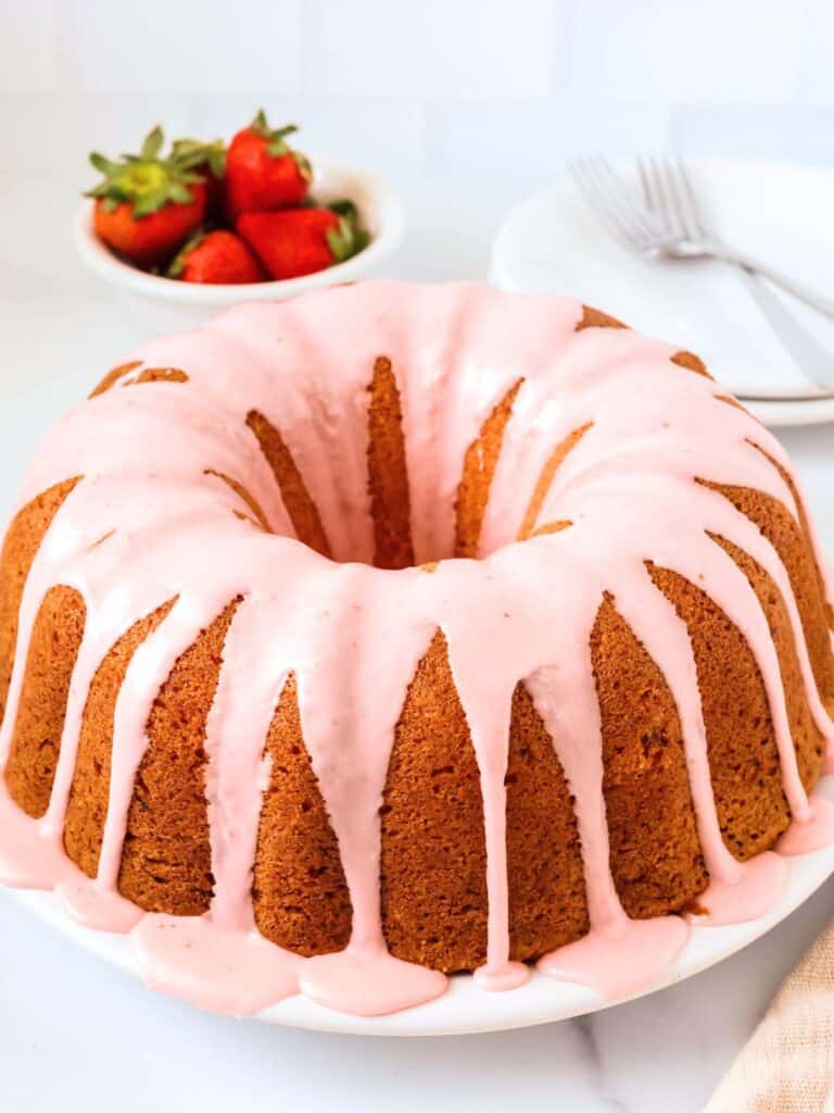 Strawberry pound cake made from a mix, topped with strawberry glaze, served on a white plate. A small bowl of fresh strawberries is in the background, along with dessert plates and forks.