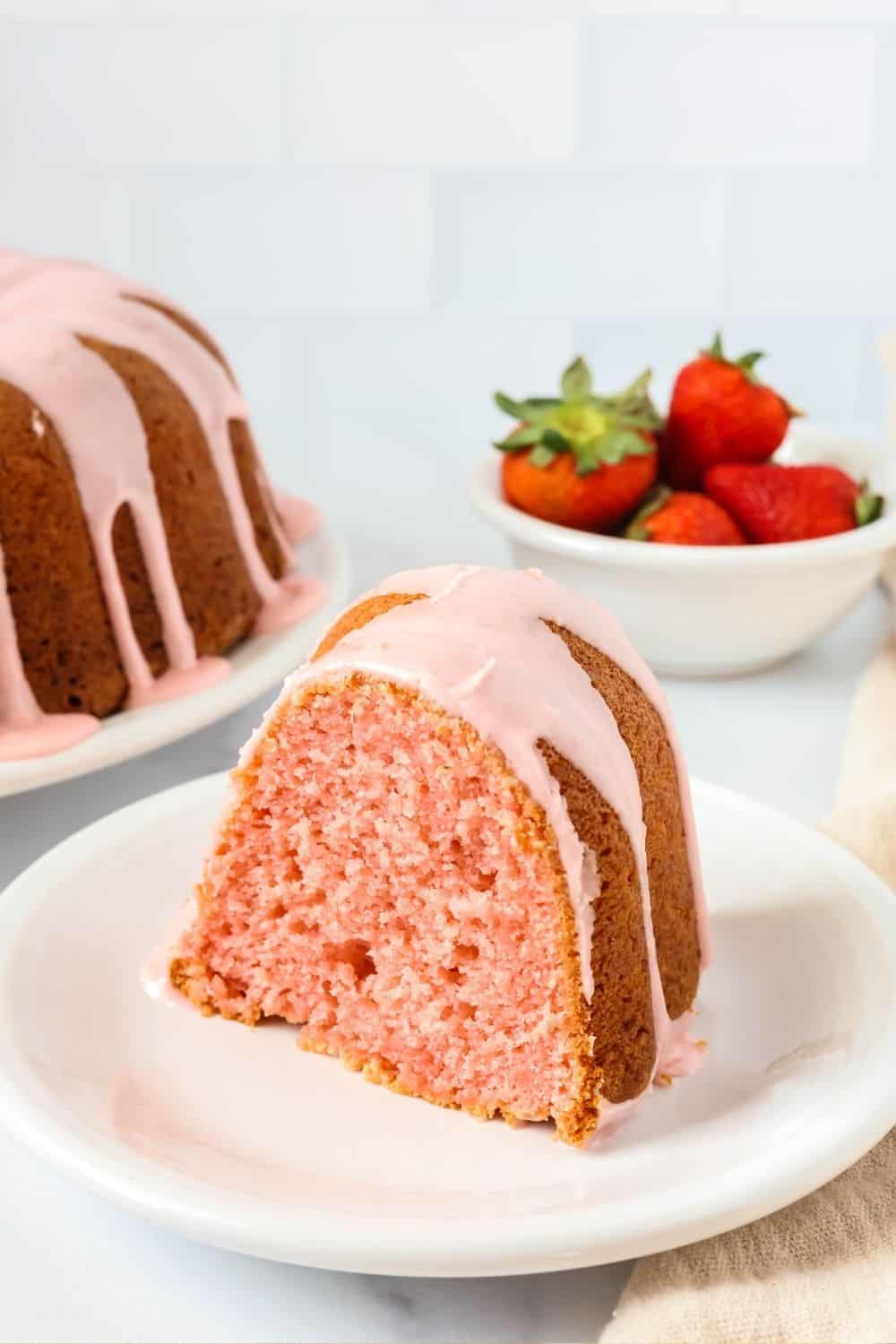 Slice of strawberry pound cake made from a mix, served on a white plate. A bowl of strawberries is in the background, along with the remainder of the bundt cake.