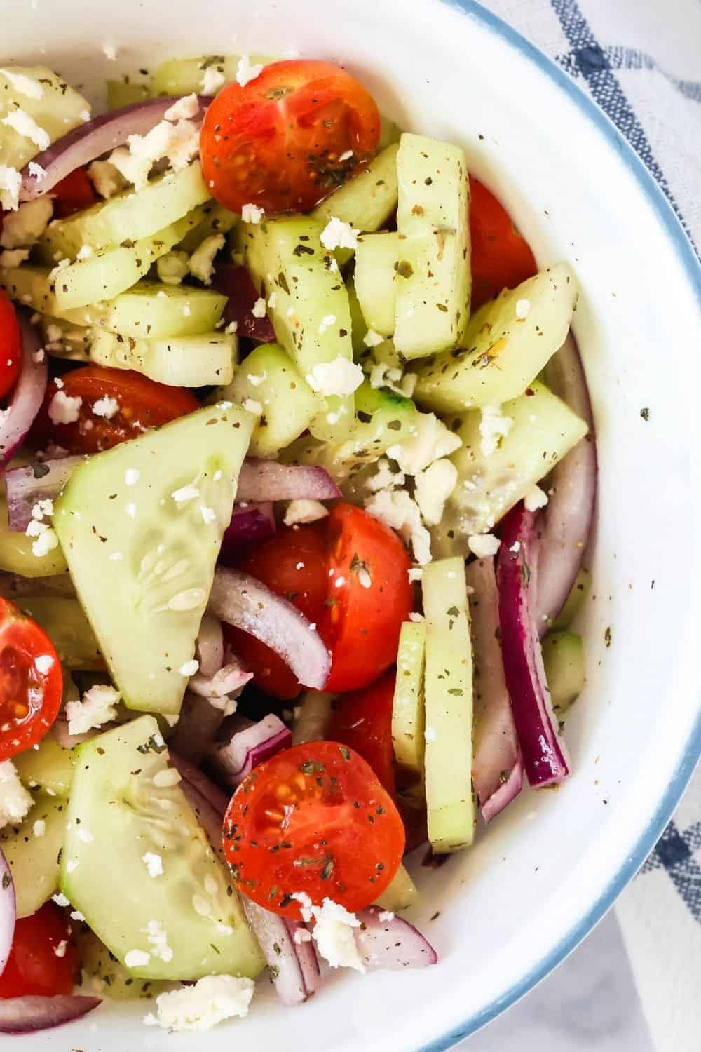 overhead close-up view of summer cucumber salad in a white bowl, complete with cucumbers, tomatoes, red onions, herbs, and feta cheese.