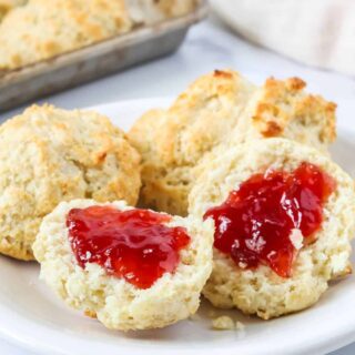 Bisquick drop biscuits served on a white plate. One biscuit is split open and topped with strawberry jam, showing the fluffy interior of the biscuit.