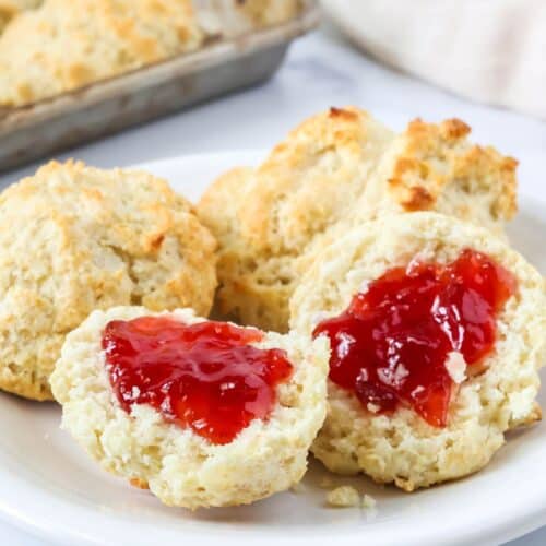 Bisquick drop biscuits served on a white plate. One biscuit is split open and topped with strawberry jam, showing the fluffy interior of the biscuit.